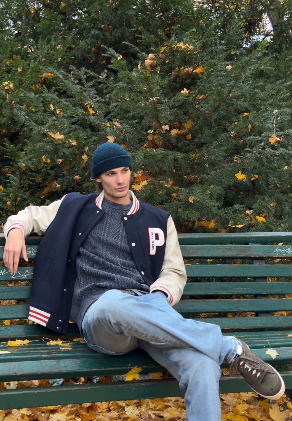 Marineblauw en crèmekleurig varsityjack met rode accenten, gestructureerde grijze trui, blauwe spijkerbroek, zittend op een groen bankje in een bosrijk gebied met herfstbladeren.