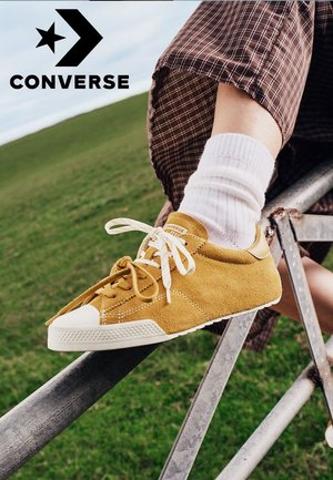 Zapatilla Converse de ante amarilla con cordones blancos y calcetines, usada por una persona sentada en una estructura metálica en un área al aire libre con césped.