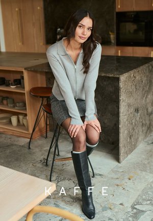 Grauer Reißverschluss-Pullover mit Kragen, kombiniert mit einem karierten Rock und kniehohen schwarzen Lederstiefeln. Auf einem Holzbarhocker sitzend.