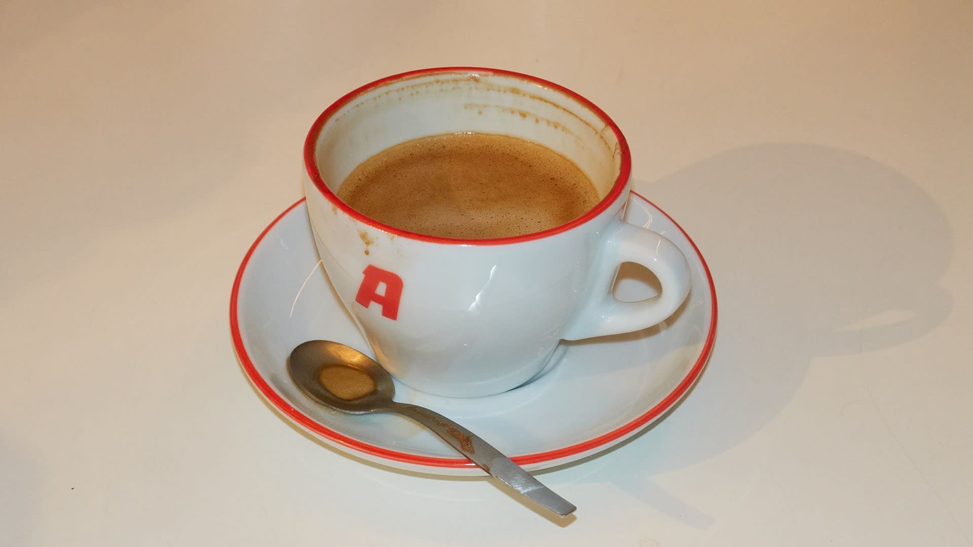 Witte keramische koffiekop met een rode rand en de letter "A," rustend op een bijpassend schoteltje, vergezeld door een zilveren lepel. Koffie is zichtbaar binnenin.