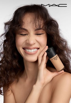 Bouteille de fond de teint MAC Studio Fix avec un capuchon noir, conçue en forme cylindrique, tenue par une personne aux cheveux bruns ondulés sur un fond neutre.