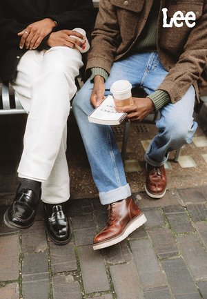 Schwarze Lederloafer neben weissen Hosen; braune Lederstiefel neben blauen Jeans; Papiertasse mit Kaffee und Buch in der Hand, auf einer Bank sitzend.