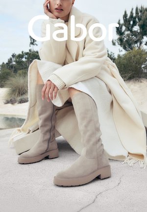 Knee-high taupe suede boots with a block heel, featuring a side seam detail and textured finish, paired with a cream coat in a natural setting.