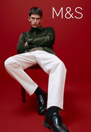 Veste en daim verte, pantalon blanc à coupe décontractée, chaussures en cuir noir et chaussettes noires. Modèle assis devant un fond rouge.