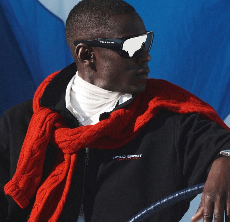 Hombre con gafas de sol Polo Sport reflectantes, chaqueta negra, suéter rojo sobre los hombros y camiseta de cuello alto blanca, sosteniendo un volante.