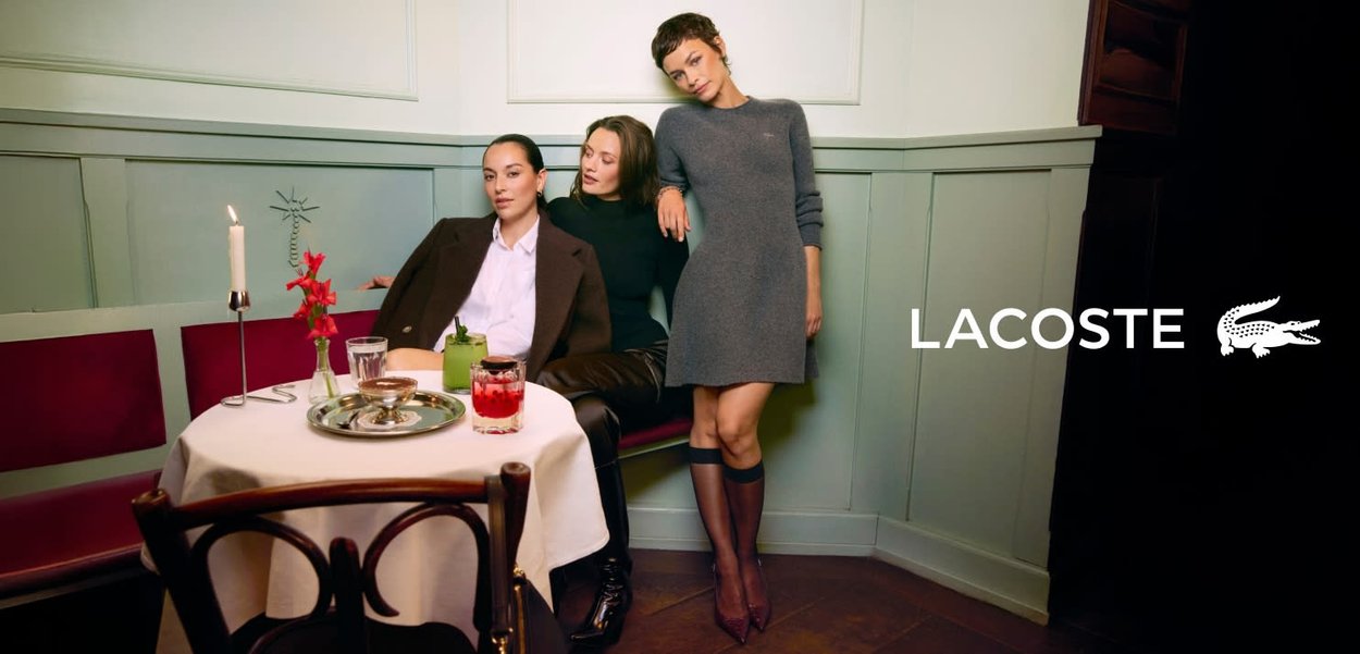 Trois femmes sont assises à une table recouverte d'une nappe blanche. Elles portent un blazer marron, un haut noir et une robe grise. Des bougies et des boissons colorées sont visibles.