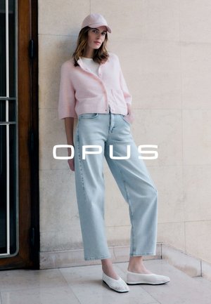 Femme portant un cardigan rose, un haut blanc, un jean bleu clair, une casquette rose et des chaussures blanches tissées sans lacets, debout contre un mur carrelé beige.