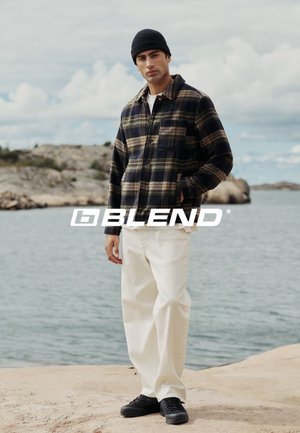 Zwart en olijfgroene geruite shirtjas, witte broek, zwarte schoenen en een beanie; staand op een rots bij het water, met een bewolkte lucht op de achtergrond.