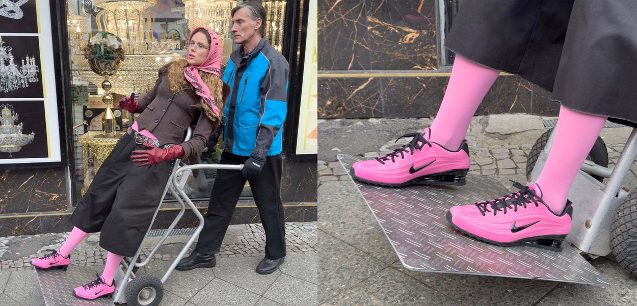 Una donna con un completo marrone, calze rosa e scarpe da ginnastica rosa è sdraiata in un carrello metallico. Un uomo con una giacca blu spinge il carrello.
