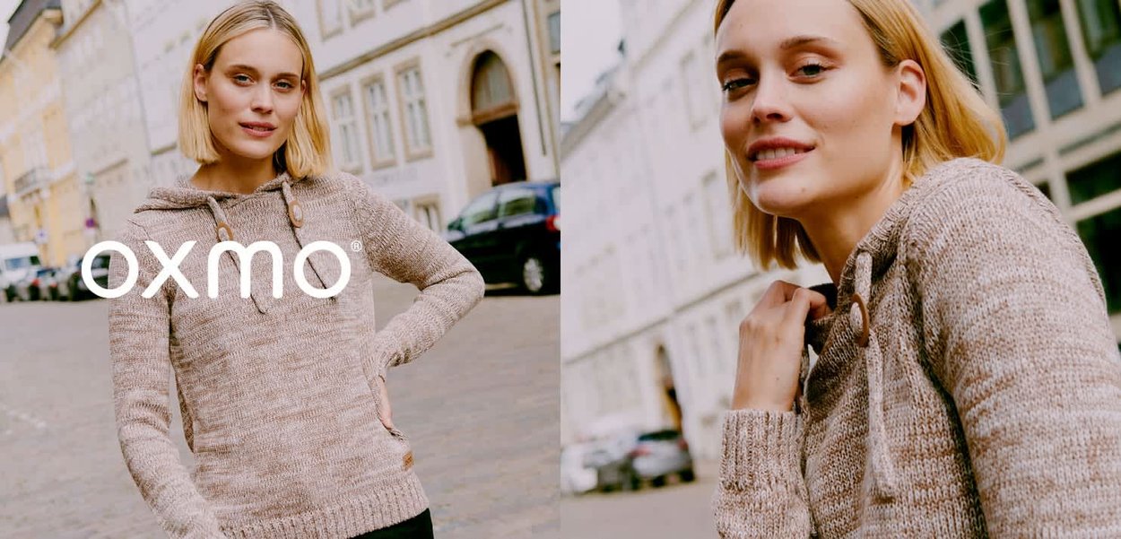 Blonde vrouw in een beige gebreide trui met knopen op de kraag, staand op een stadsstraat met historische gebouwen en geparkeerde auto's.