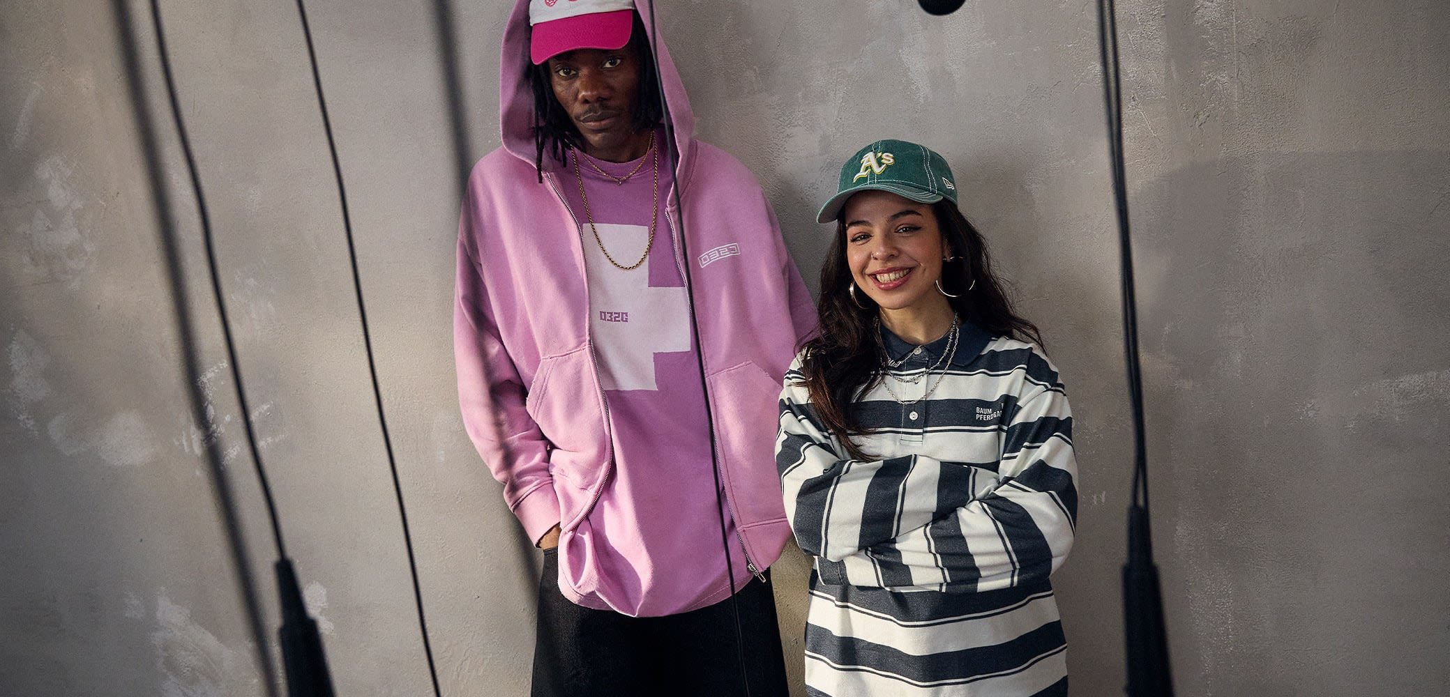 Due persone indossano abiti casual: una in un hoodie rosa con una grafica e una maglietta viola, l'altra in una maglietta a maniche lunghe a righe e un cappello verde.