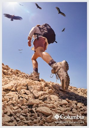 Wanderschuhe aus strapazierfähigem grauem Material steigen über felsiges Terrain, während ein Rucksack auf dem Rücken getragen wird. Klarer blauer Himmel und Vögel darüber.