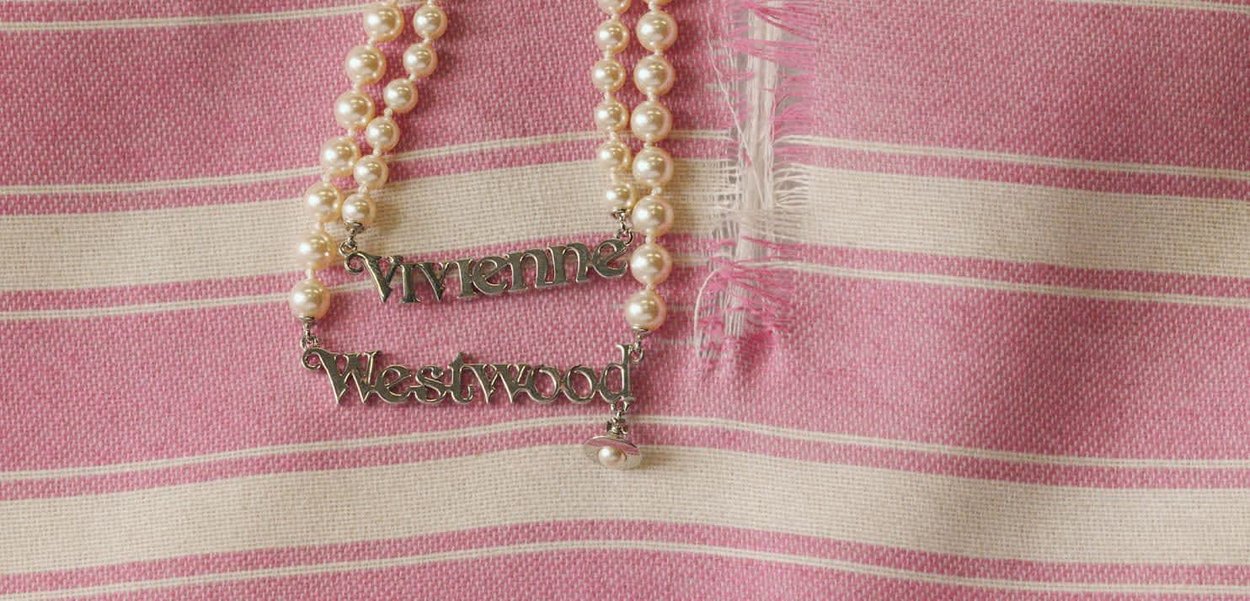 Collier couleur argent présentant deux plaques nominatives "Vivienne" et "Westwood" suspendues entre des brins de perles blanches crémeuses sur un fond rayé rose.