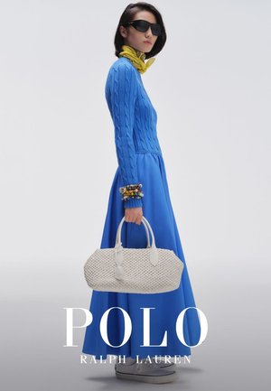 Mujer con suéter azul de punto trenzado y falda, pañuelo amarillo en el cuello, gafas de sol oscuras, sosteniendo un bolso blanco tejido, posando frente a un fondo gris.