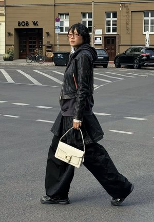 Schwarze Lederjacke über einem dunklen Hoodie, kombiniert mit einem schwarzen, strukturierten Rock und weiten Hosen. Weiße Handtasche mit einem goldenen Verschluss.