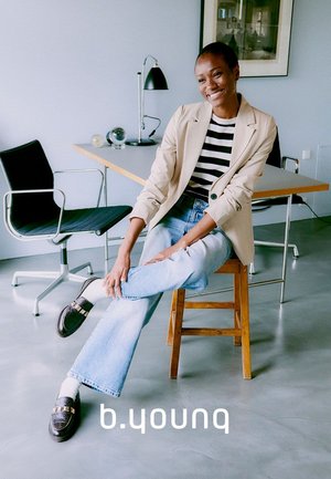 Light beige blazer over a black and white striped shirt, paired with light blue flared jeans and dark loafers with metallic accents, seated on a stool.