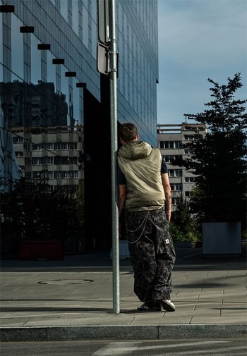 Persoon leunend tegen een metalen paal, gekleed in een lichtgroene mouwloze jas en donkere camouflagebroek, staande op een bestrate stoep in de stad.