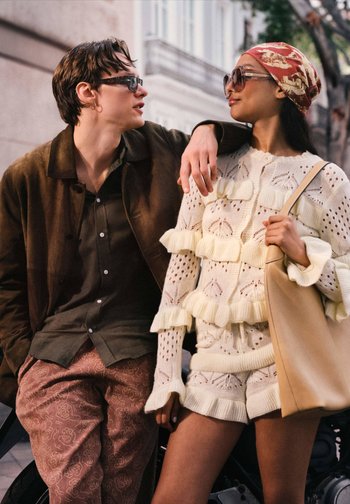 Knitani pulover s volanima u krem boji, uparen s kratkim hlačama u istoj boji. Momak u smešnom zimskoj jakni od smešnog baršuna i sa uzorkovanim hlačama, oboje nose sunčane naočale.