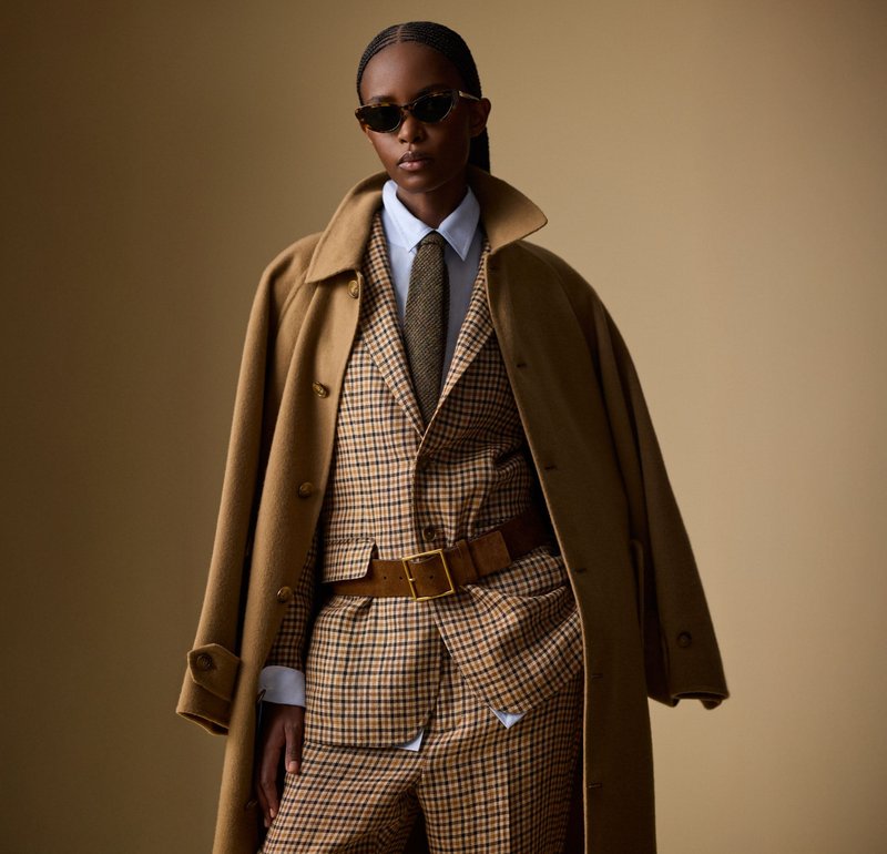 Persona con un traje de cuadros marrón y una corbata a juego, camisa blanca, abrigo de camello oversize colgado sobre los hombros y gafas de sol oscuras, todo esto frente a un fondo beige.