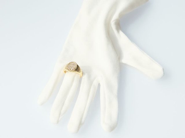Λευκό ύφασμα γάντι κρατά έναν χρυσό μεταλλικό δακτύλιο διακοσμημένο με διαφανείς πέτρες, που παρουσιάζει στρογγυλό σχήμα με πολύπλοκες λεπτομέρειες.