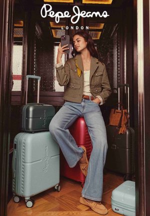 Joven mujer con chaqueta informal y vaqueros sentada sobre una maleta roja, tomando una selfie con su smartphone, rodeada de varias piezas de equipaje de Pepe Jeans.