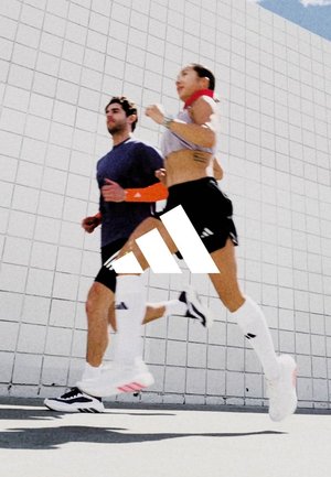 Zwei Personen laufen draußen vor einer weißen gefliesten Wand, tragen Sportbekleidung und weiße knielange Socken mit Markensneakern.