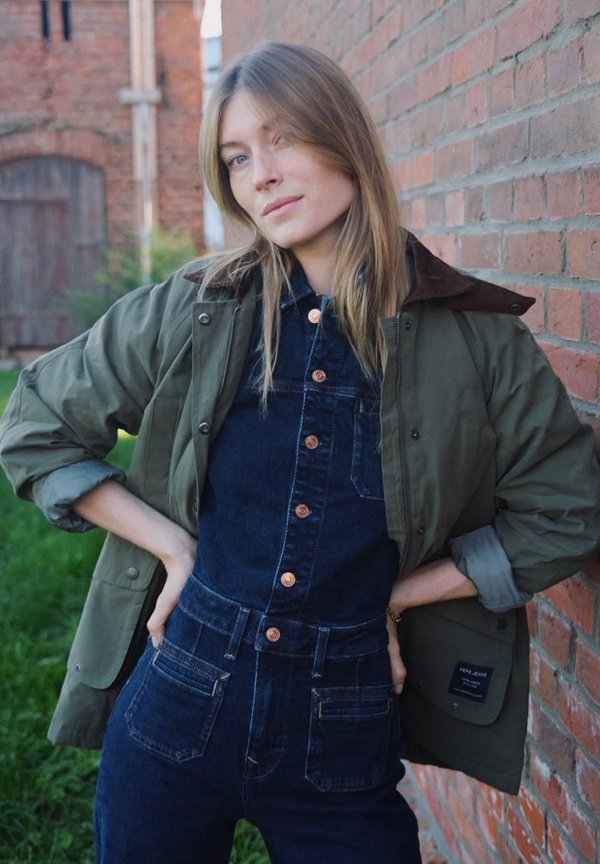 Green jacket with brown collar over a dark denim jumpsuit, featuring copper buttons and multiple pockets, against a brick wall background.
