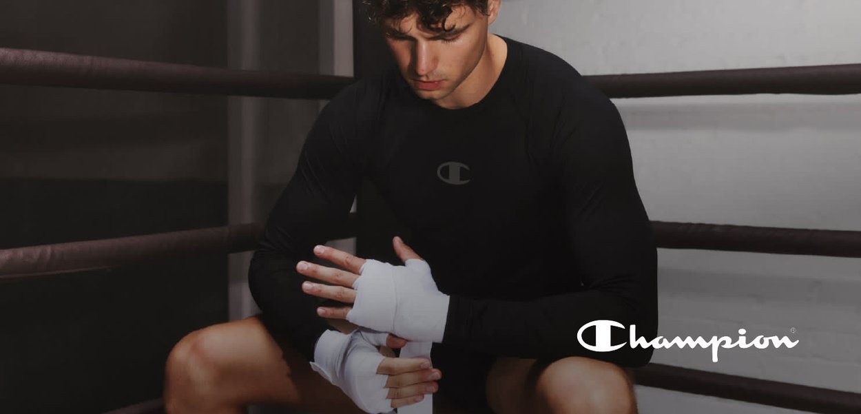 Camisa atlética de manga larga negra con logo, vendajes blancos en las manos, sentado en un ring de boxeo con acolchado marrón. Enfoque en las manos y el diseño de la camisa.