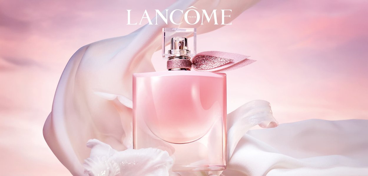 Pink perfume bottle with a rounded base, clear cap, and glittery accent. Features a pink satin ribbon and is set against soft, flowing fabric.