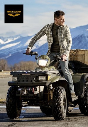 Homme en chemise à carreaux et jean debout sur un VTT Polaris vert avec des montagnes enneigées en arrière-plan sous un ciel dégagé.