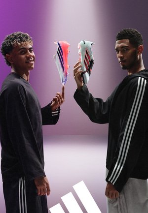 Dwóch mężczyzn w czarnych strojach Adidas trzyma po jednej korku piłkarskiej, jedna różowa i jedna biała, na fioletowym gradientowym tle z białym logo Adidas.