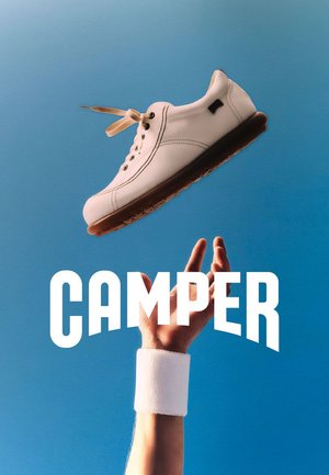 Mano con una pulsera blanca lanzando una zapatilla blanca con suela marrón contra un cielo azul despejado, con el texto destacado "CAMPER" debajo.