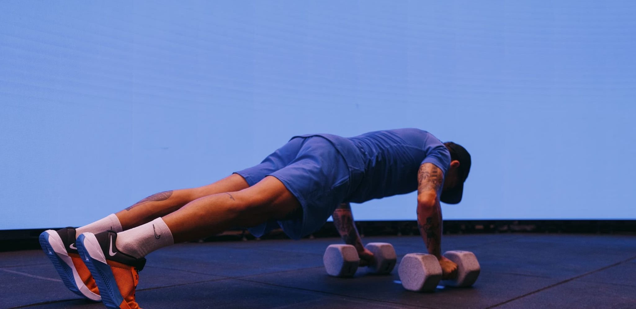 Gray hexagonal dumbbells on a black gym mat. A person in a blue athletic outfit performs push-ups, with an abstract blue background.