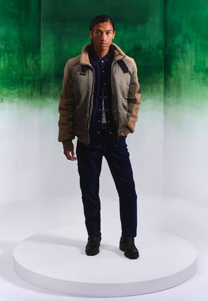 Gevoerde olijfgroene bomberjack met beige schapenvacht mouwen, denim overhemd, donkere jeans en zwarte laarzen, geposeerd op een ronde plaat tegen een witte achtergrond.