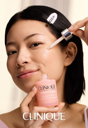 Femme aux cheveux foncés utilisant un compte-gouttes pour appliquer le sérum Clinique Moisture Surge sur sa joue, souriant doucement avec une pince à cheveux en place.