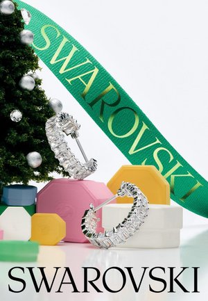 Boucles d'oreilles créoles en argent avec cristaux clairs, un ruban vert avec l'inscription "SWAROVSKI" et des boîtes cadeaux hexagonales colorées sur fond blanc.