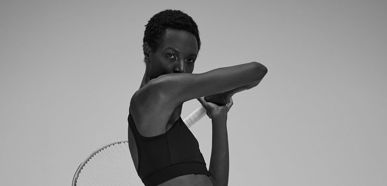 Reggiseno sportivo nero, texture liscia, design aderente, con un colore solido. Persona che tiene una racchetta da tennis, postura atletica su uno sfondo grigio.