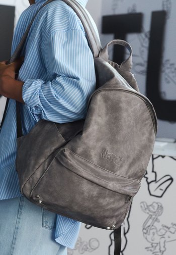 Grauer Leder-Rucksack in runder Form, mit sichtbaren Nähten, Fronttasche und silbernem Zubehör. Über der Schulter getragen mit einem blau gestreiften Hemd.