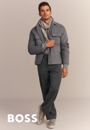 Graue Wollmischjacke mit zwei frontalen Taschen, über einem hellgrauen Pullover getragen. Hochgeschnittene dunkle Hose und beige Sneakers runden das Outfit ab.