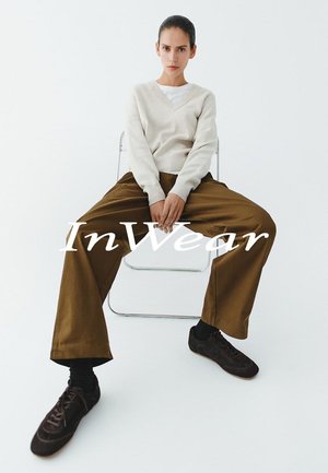 Cremefarbener V-Ausschnitt-Pullover kombiniert mit lockeren braunen Hosen. Die Person trägt schwarze Socken und dunkelbraune Schnürschuhe und sitzt auf einem transparenten Stuhl.