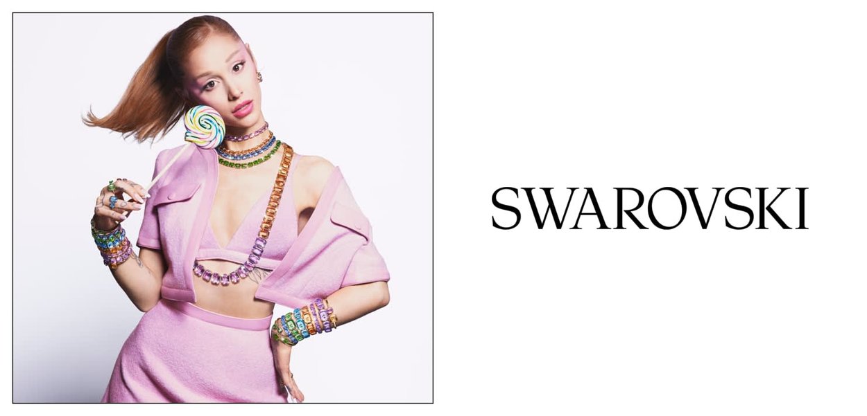 Giovane donna con abito rosa che indossa gioielli di cristallo colorati e tiene un grande lecca-lecca a spirale accanto al logo Swarovski su sfondo bianco.