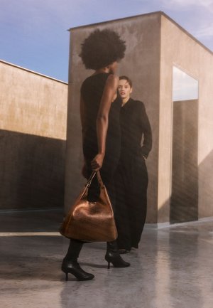 Black sleeveless dress, woman carrying a textured brown leather bag, wearing black heels. Concrete background with a glass mirror reflects light.