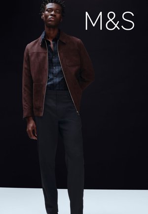 Veste en daim marron avec fermeture éclair à l'avant, portée sur une chemise à motif bleu marine, associée à un pantalon foncé. Design simple sur un fond neutre.