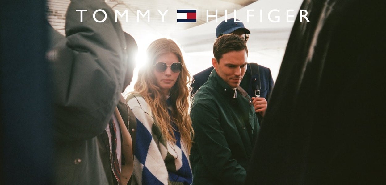 Imagen promocional de Tommy Hilfiger que presenta a individuos con una mezcla de ropa oscura y con estampados. La luz del sol crea un suave resplandor en el fondo.