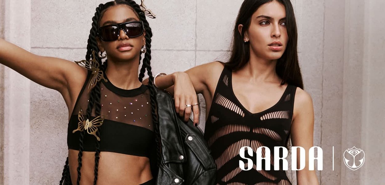 Dos mujeres posan frente a una pared de hormigón, una con gafas de sol y accesorios de mariposas, la otra con una camiseta de malla negra con el logo SARDA.