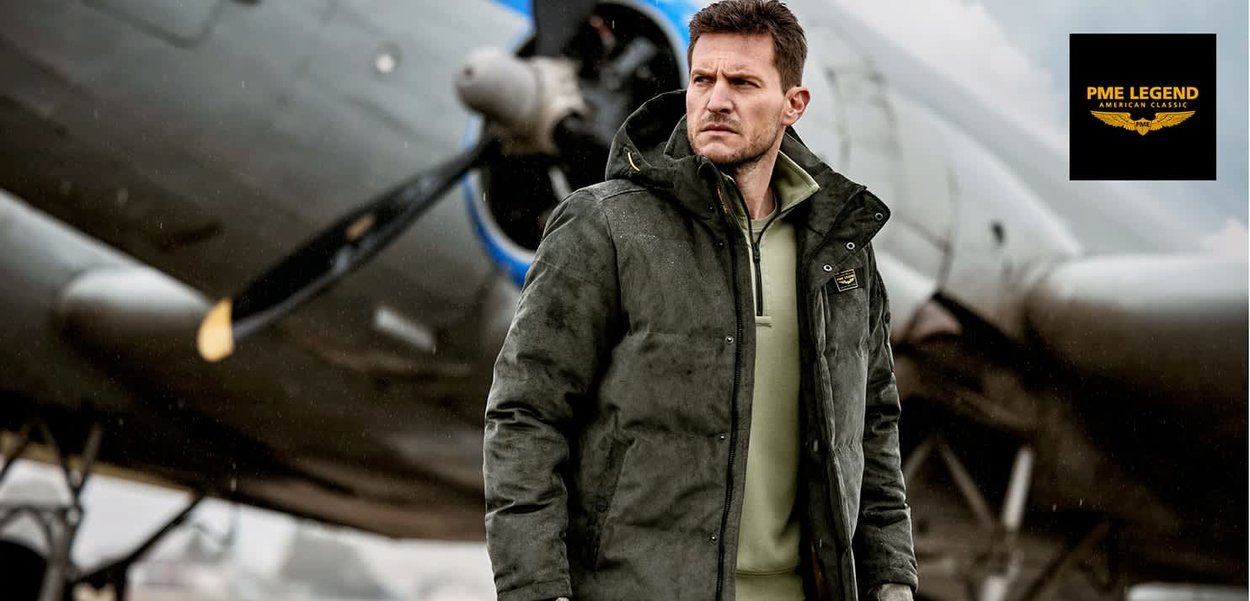 Abrigo exterior texturizado color verde oliva con cremallera frontal y cuello grande, superpuesto a un suéter verde claro, con un fondo de una aeronave vintage.