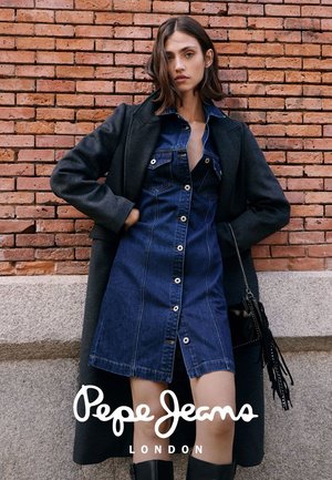 Mujer con abrigo gris oscuro sobre vestido vaquero abotonado, botas negras, sosteniendo bolso negro con flecos, de pie frente a una pared de ladrillos rojos.