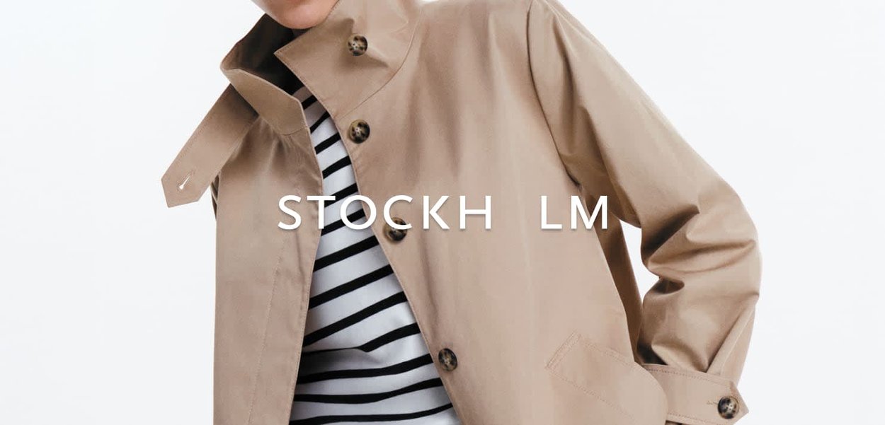 Persoon draagt een beige trenchcoat over een zwart-wit gestreept shirt, tegen een effen witte achtergrond.