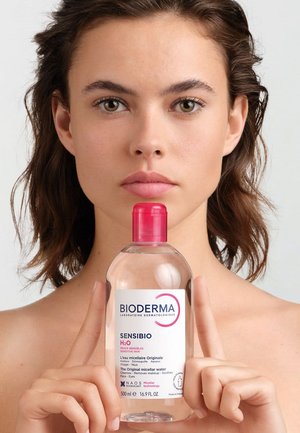 Junge Frau mit natürlicher Haut hält eine klare Bioderma Sensibio Mizellenwasserflasche mit rotem Deckel, die mittig unter ihrem Gesicht positioniert ist.