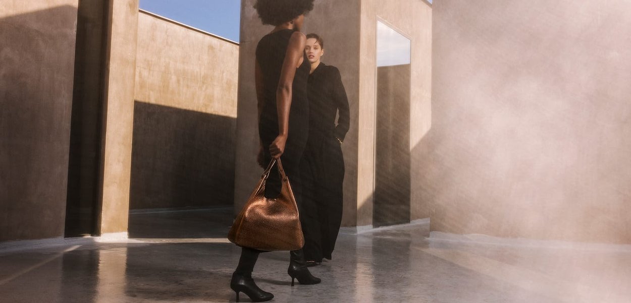 Brown textured handbag with a hobo design. Two individuals in black outfits standing in a modern, minimalist space with soft light.